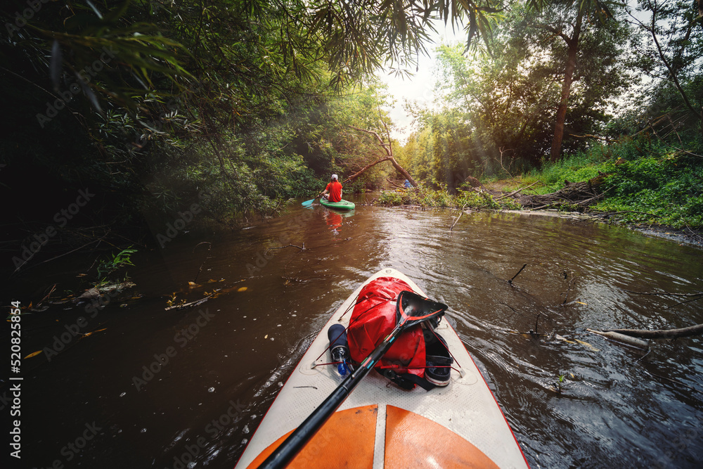 Fototapeta premium View from the first person. A trip on a SAP board. A narrow river in the middle of the forest, an extreme. Red waterproof bag. Tours and tourism, active recreation. the sun's rays are shining
