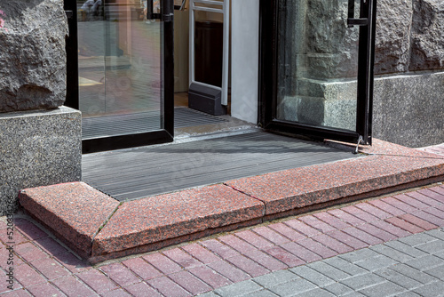 entrance with granite threshold to store from pedestrian sidewalk from plink on foot mat through glass door with black frame, rough stone facade of exterior of building close-up in sunny day, nobody.