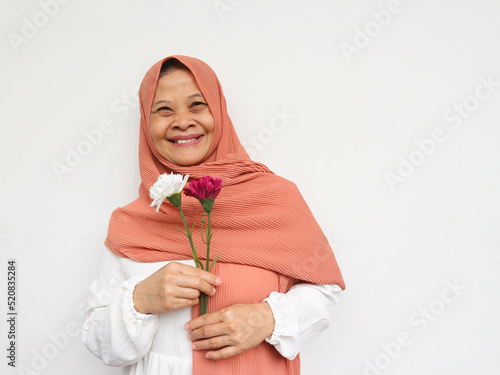 Beautiful face of happy mature middle-aged woman in hijab. Elderly woman in turban, healthy cheerful smiling, looking at camera while holding flowers and posing at white background