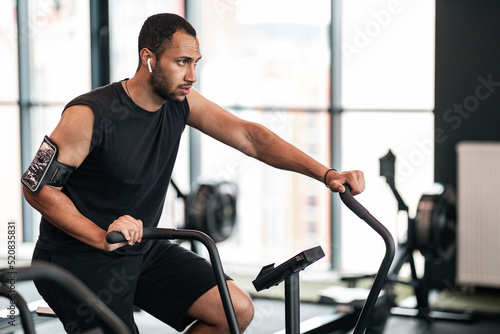 Sporty Young Black Man Using Elliptical Bike Machine While Training At Gym