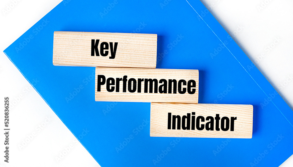 There is a blue notebook on a light gray background. Above are three wooden blocks with the words Key Performance Indicator There is a blue notebook on a light gray background. Above are three wooden blocks with the words Key Performance Indicator