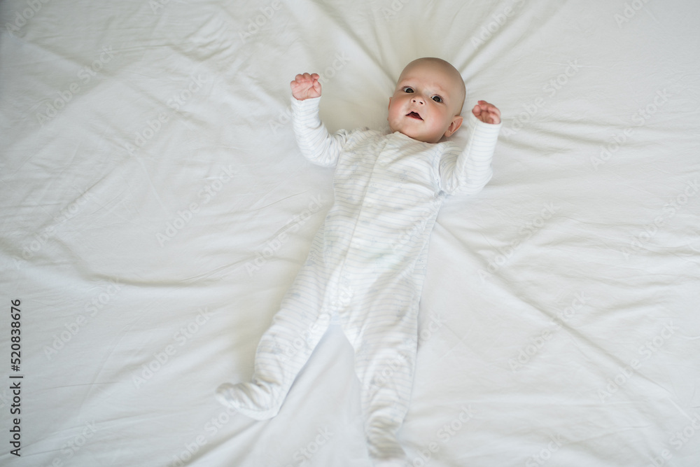 Happy baby in white overalls lying on the bed, smiling infant in white clothes, six month old baby Happy baby in white overalls lying on the bed, smiling infant in white clothes, six month old baby