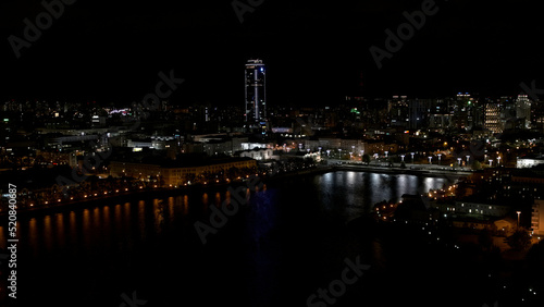 The view from the drone.Stock footage.A beautiful city at night located next to the river and beautiful high-rise new buildings and offices and with a calm illuminated road