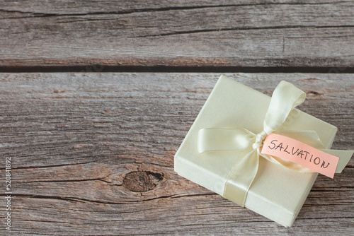 White gift box with ribbon and handwritten word "salvation" on a note placed on a rustic wooden table with copy space. Top view. Christian biblical concept of the blessing of eternal life.