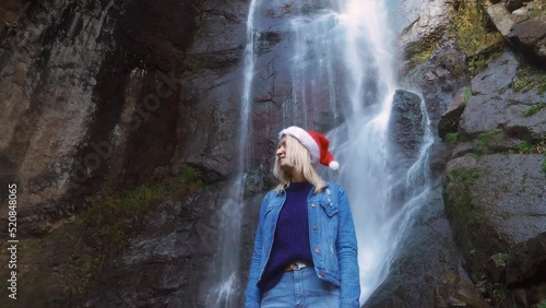 A young blonde in a Santa Claus hat stands at Makhuntseti Waterfall in Georgia. female traveler celebrates a walk on Christmas Day. Christmas nature. Christmas Eve. She looks and enjoys nature around. photo