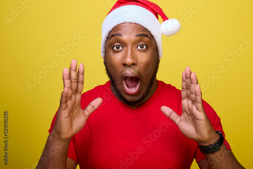 Shocked young African-American Santa Claus in a red T-shirt and a Christmas hat, shouting with a hand gesture at his mouth, isolated on a yellow background. Concept Happy New Year, Merry Christmas © Aleksandr