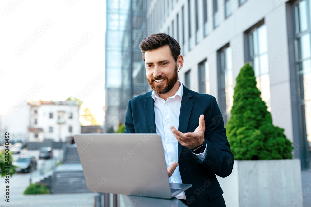 Happy millennial attractive caucasian guy with beard in suit chatting on laptop, has video call on balcony Happy millennial attractive caucasian guy with beard in suit chatting on laptop, has video call on balcony
