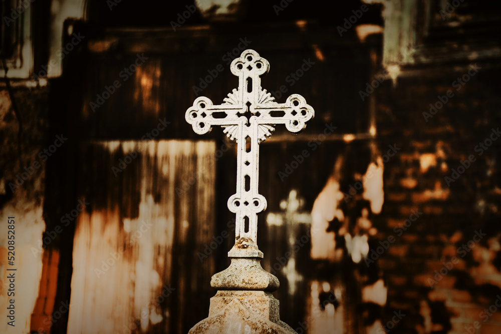 A white Christian cross stands on a pedestal above the grave in the cemetery against the background of the crypt, illuminated by the rays of the setting sun. Religion and death.