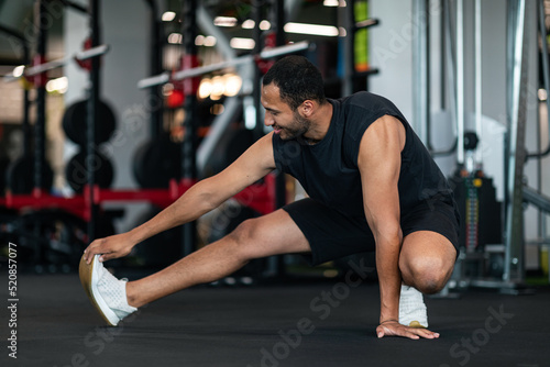 Sporty Young Black Man Stretching Leg Muscles While Training At Gym