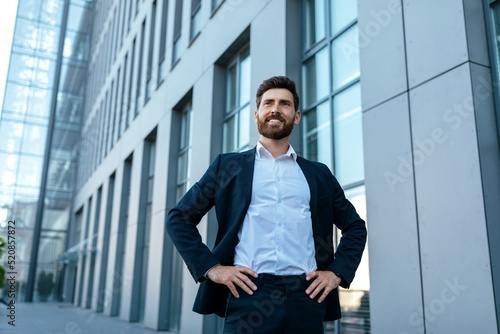 Cheerful confident handsome millennial businessman with beard in suit stands near modern office building