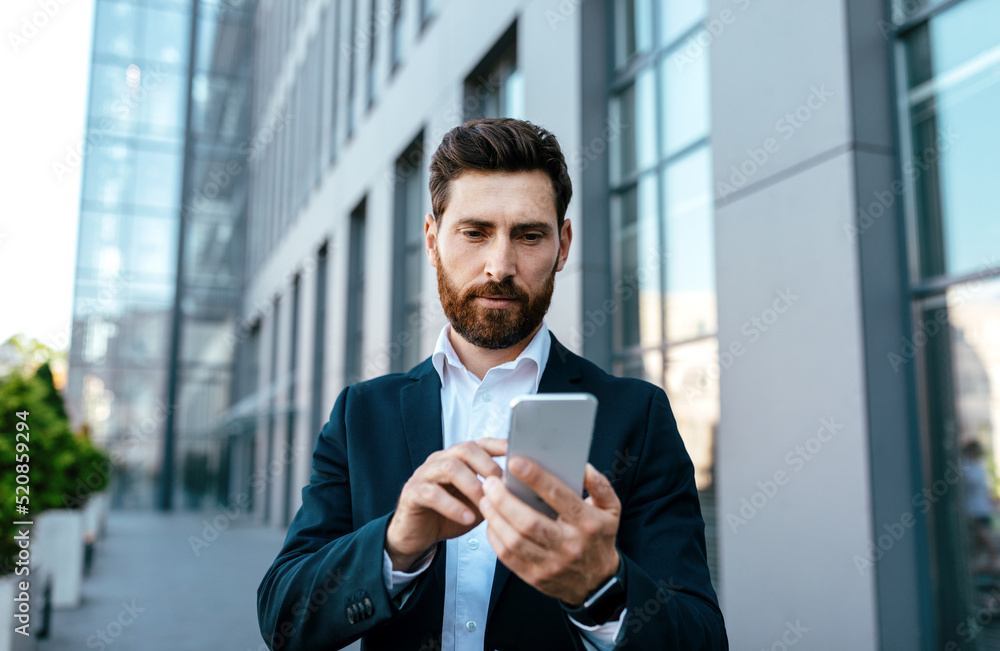 Serious confident millennial caucasian businessman with beard in suit typing on smartphone near office building Serious confident millennial caucasian businessman with beard in suit typing on smartphone near office building