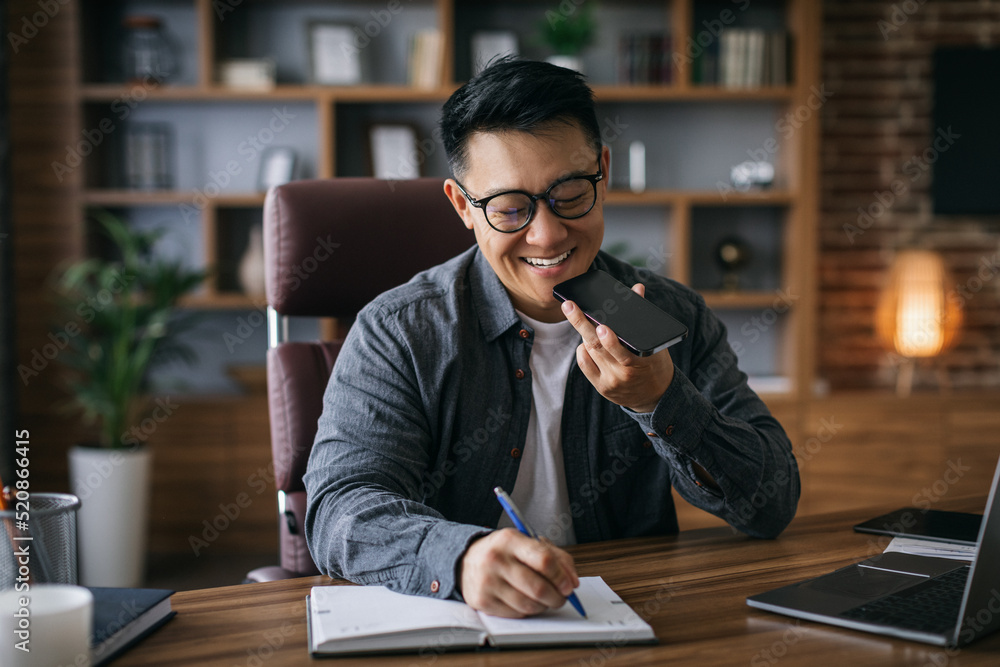 Smiling mature asian businessman in glasses talking on smartphone and making notes at workplace in home