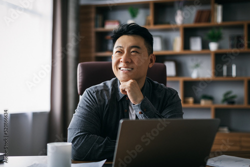 Smiling pensive adult asian businessman inspired by idea at laptop to workplace in home office interior, free space