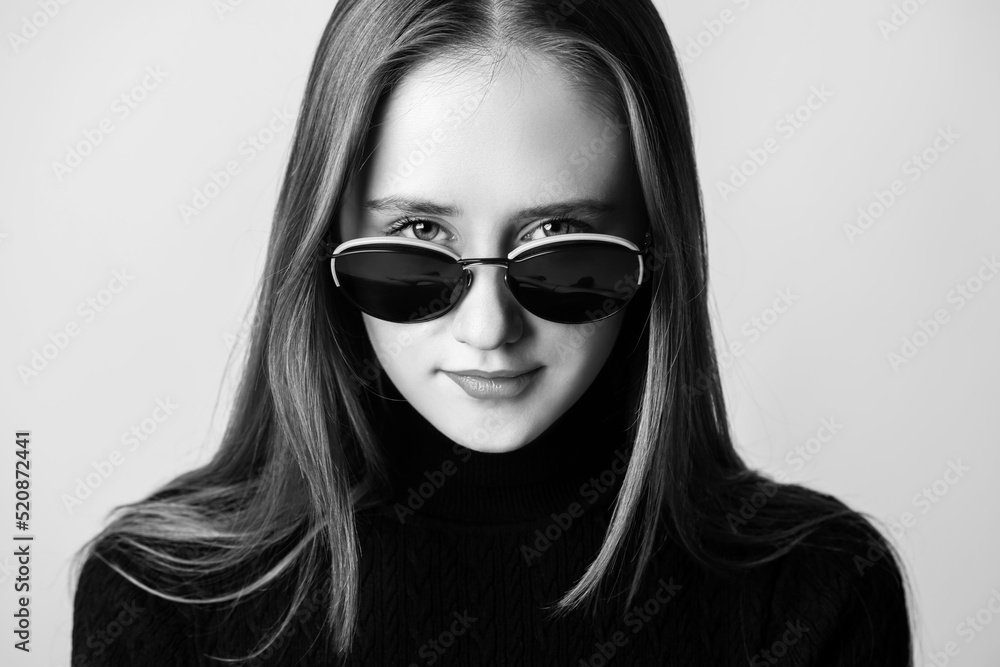 Make-up, beauty, style and fashion concept. Close-up woman portrait looking through sunglasses direct to camera and wearing black long neck sweater in grey studio background. Black and white image Make-up, beauty, style and fashion concept. Close-up woman portrait looking through sunglasses direct to camera and wearing black long neck sweater in grey studio background. Black and white image