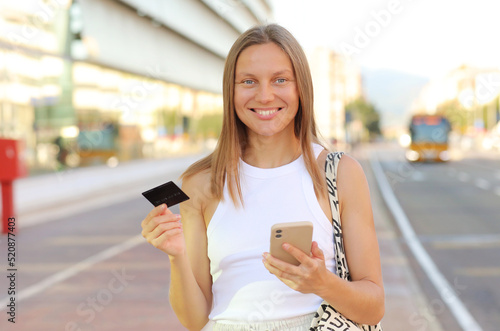 Beautiful smiling young woman consumer with credit card and smartphone looks in camera. people shopping via online application media concept. shopping mall on summer day background with copy space.