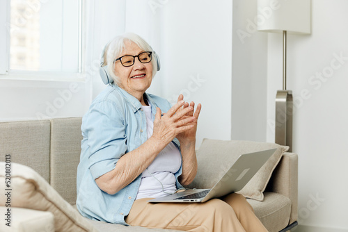 a happy elderly lady with gray hair is sitting on a cozy sofa talking via video link via laptop and headphones and smiling broadly holding her hands in a relaxed gesture © Tatiana