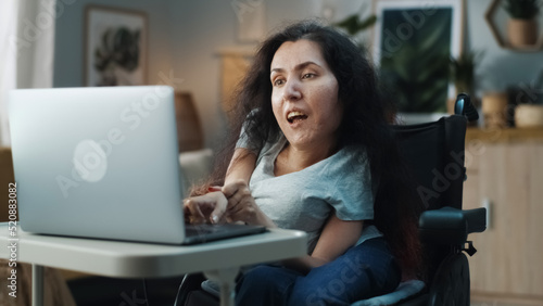 Fotografie Woman with disability sitting in a wheelchair at home in a cozy room recording a