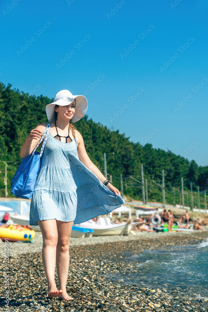 young beautiful woman traveler in a blue summer sundress, sun hat and with a beach bag walks along the seashore during a summer vacation