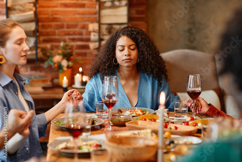 Portrait of young black woman holding hands with friends and saying grace at dinner party in cozy setting  eyes closed