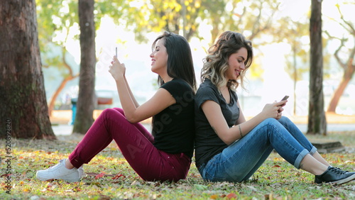 Friends together sharing internet media content together outdoors Girlfriends seated outside at the park using smartphones