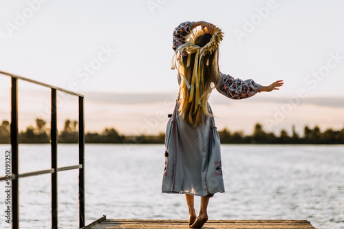 A beautiful, happy Ukrainian girl dances on the pier, rejoicing in freedom, against the background of water, a river, in an embroidered shirt, a wreath, barefoot at sunset. Photography, Ukraine.