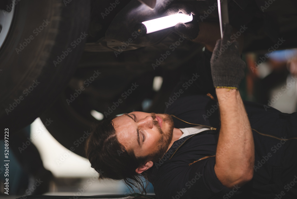 Young auto and car mechanic with beard and wearing uniform lying on floor while repairing car automobile engine from bottom in garage Young auto and car mechanic with beard and wearing uniform lying on floor while repairing car automobile engine from bottom in garage