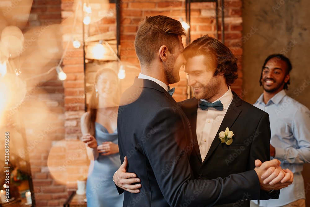 © Seventyfour - Dreamy portrait of young gay couple dancing together during wedding reception with lens flare