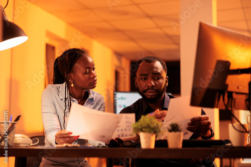 African american business colleagues doing teamwork collaboration to do presentation report  looking at data on computer and papers. Working together with paperwork in office during sunset.