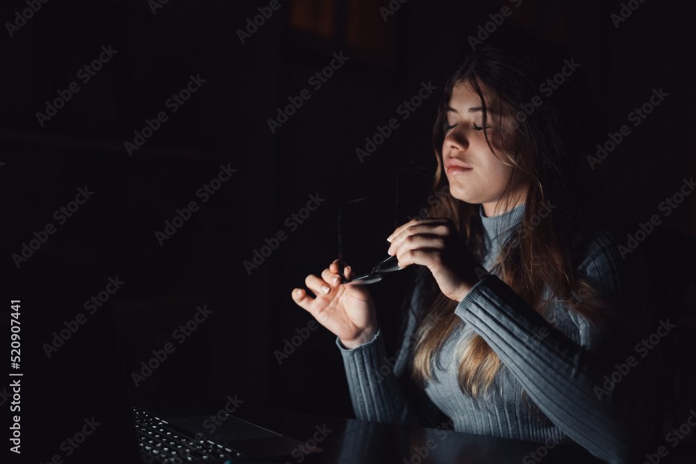 One tired and exhausted young woman taking off her eye glasses after a long time working at night at home in the dark. Caucasian girl stressed and feeling bad working with headache. One tired and exhausted young woman taking off her eye glasses after a long time working at night at home in the dark. Caucasian girl stressed and feeling bad working with headache.