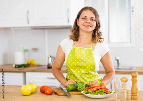 Positive woman wearing apron, standing at kitchen table while preparing salad.