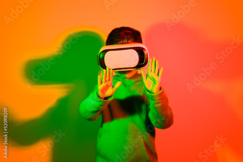 little boy standing in front of a neon colored background wearing virtual reality goggles with his hands stretched forward pretending to touch something. High quality photo