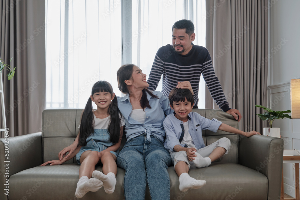 Asian Thai family, adult dad, mum with little children are happy home living, looking together, smiling and leisure on sofa in house's white room, lovely weekend and well-being domestic lifestyle.