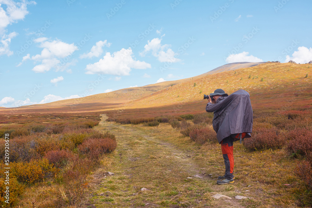 Alone traveler in raincoat with large backpack photographs nature among fading autumn vegetations on high mountain plateau. Backpacker with photo camera shoots mountain landscape in changeable weather Alone traveler in raincoat with large backpack photographs nature among fading autumn vegetations on high mountain plateau. Backpacker with photo camera shoots mountain landscape in changeable weather