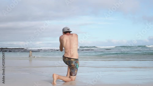 Shirtless Male Tourist Kneeling On Tortuga Bay Beach Taking Photograph Of Flying Brown Pelican With Telephotos Lens. Pan Left. Slow Motion photo