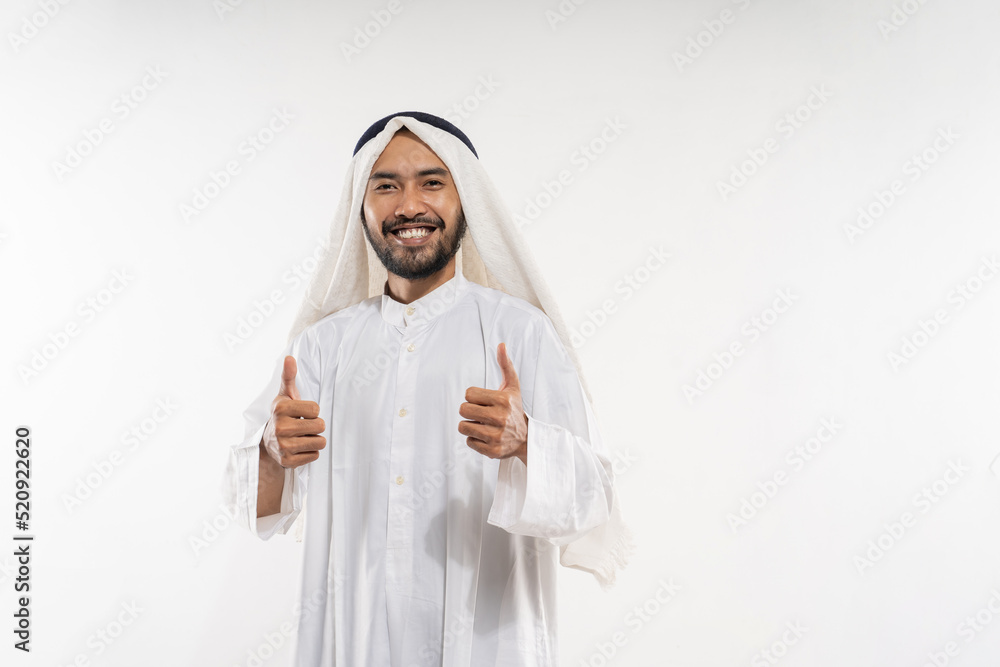 an arabian young man in a turban smiling while standing looking at the camera with thumbs up while talking against a plain background