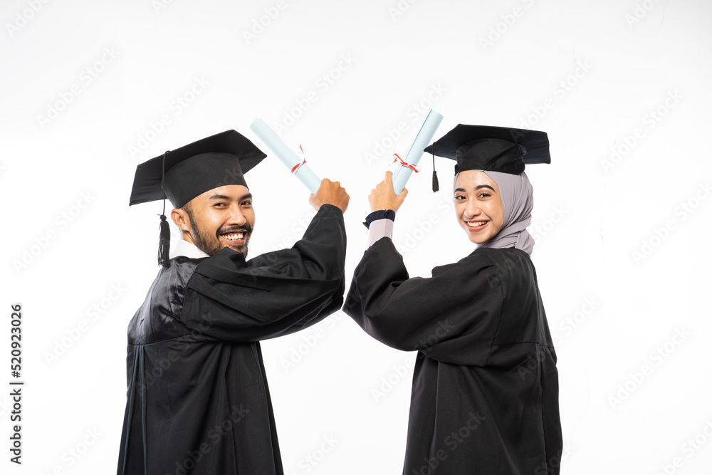 Two graduate students wearing togas holding up certificate while looking back on isolated background Two graduate students wearing togas holding up certificate while looking back on isolated background