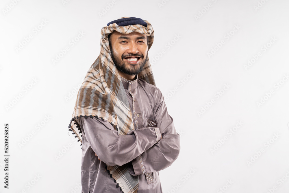 an arabian young man in a turban standing smiling with crossed hands on a plain background