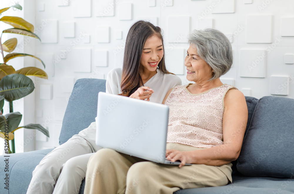Asian cheerful old senior healthy gray hair female pensioner mother sitting smiling with young daughter on cozy sofa in living room using laptop notebook computer browsing shopping online together Asian cheerful old senior healthy gray hair female pensioner mother sitting smiling with young daughter on cozy sofa in living room using laptop notebook computer browsing shopping online together