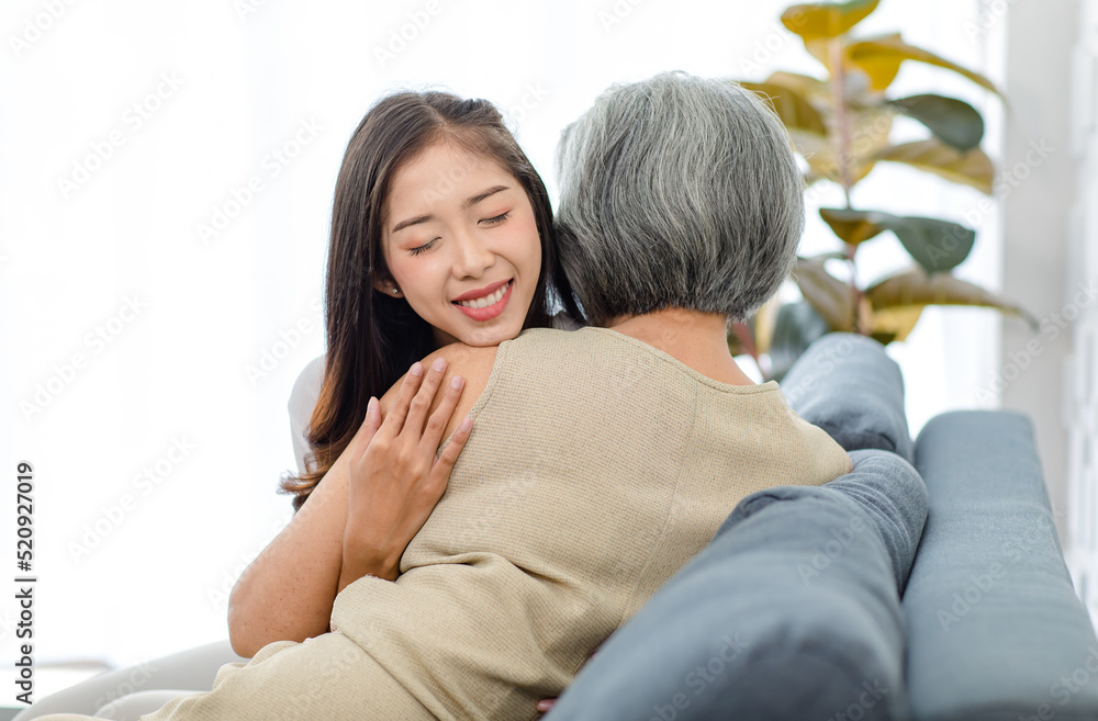 Asian cheerful young beautiful daughter sitting smiling hugging embracing showing affection love comforting bonding with old senior grey hair pensioner mother on cozy sofa in living room at home