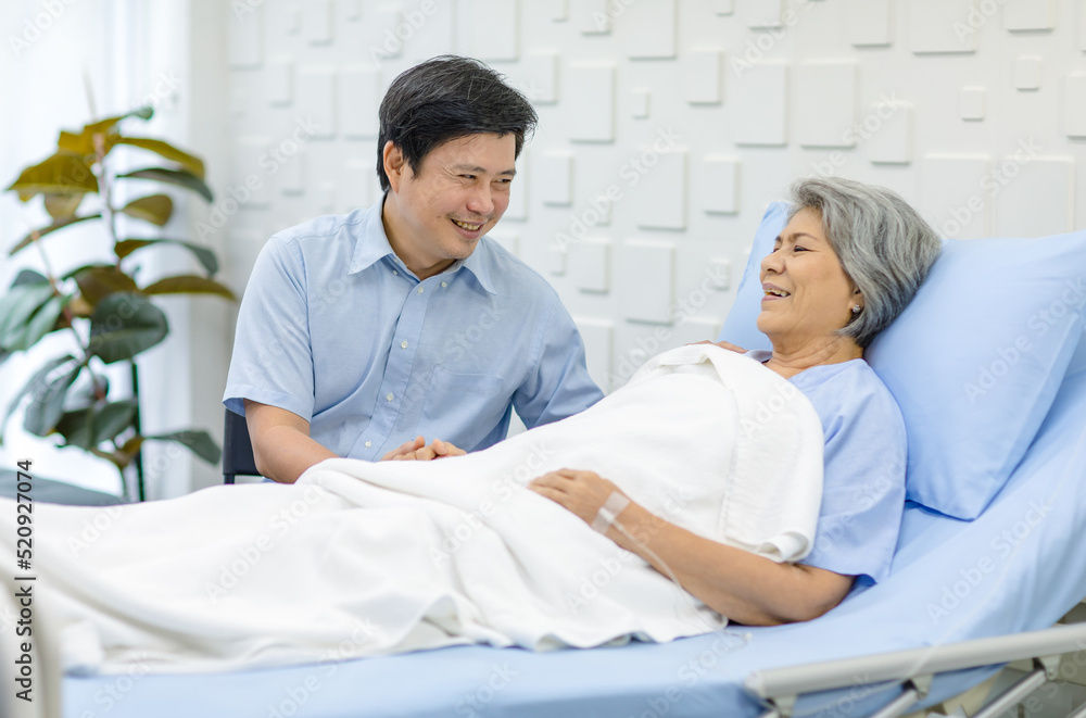 Obraz premium Asian middle aged male son visiting supporting comforting holding hands old senior sick unhealthy pensioner mother patient in blue hospital uniform laying down receiving saline solution drip on bed