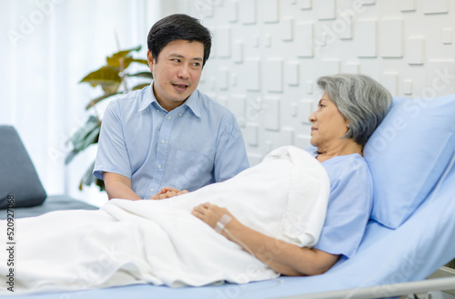 Asian middle aged male son visiting supporting comforting holding hands old senior sick unhealthy pensioner mother patient in blue hospital uniform laying down receiving saline solution drip on bed