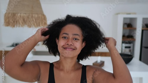 A positive African American woman with thick black wavy hair looks into the frame and straightens her Afra hairstyle. A mixed race woman smiles sweetly into the frame. Beautiful black woman model.