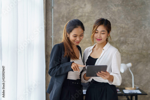 Two beautiful Asian business women stand and talk about work using a tablet in the office.