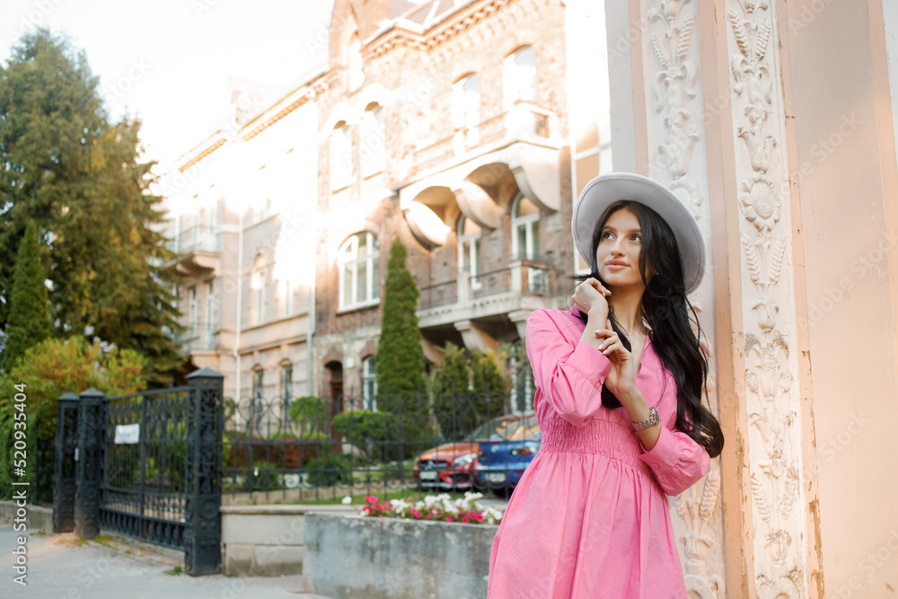 Fototapeta premium Street style, elegant woman wearing trendy pink dress, white color hat, posing in street of city.