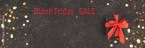 Black gift box with red bow on black concrete background with gold bokeh. Banner with copy space. Black Friday SALE.