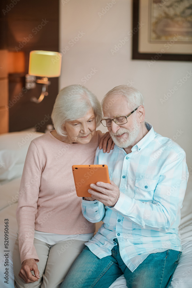 Senior couple sitting on a bed and watching something on a talet Senior couple sitting on a bed and watching something on a talet