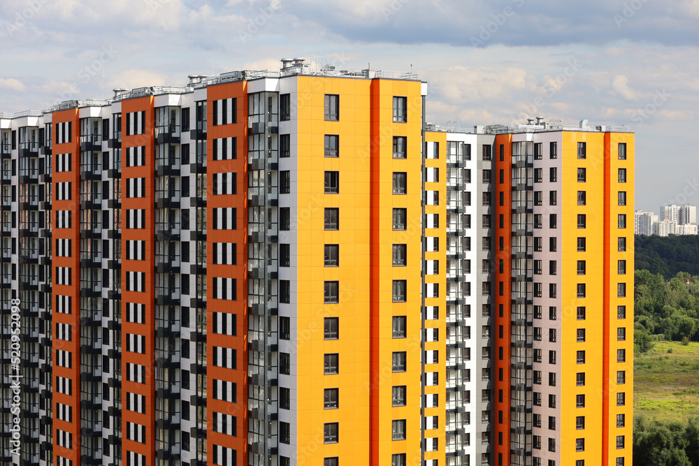 New residential buildings with orange and yellow cladding on background of green park and sky with white clouds. House development, high-rise construction in ecologically clean area New residential buildings with orange and yellow cladding on background of green park and sky with white clouds. House development, high-rise construction in ecologically clean area