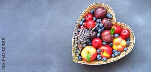 Red apples, blueberry and plums in straw basket made in the shape of heart on dark shade of blue background. Creative copy space for seasonal projects and basis designs. 