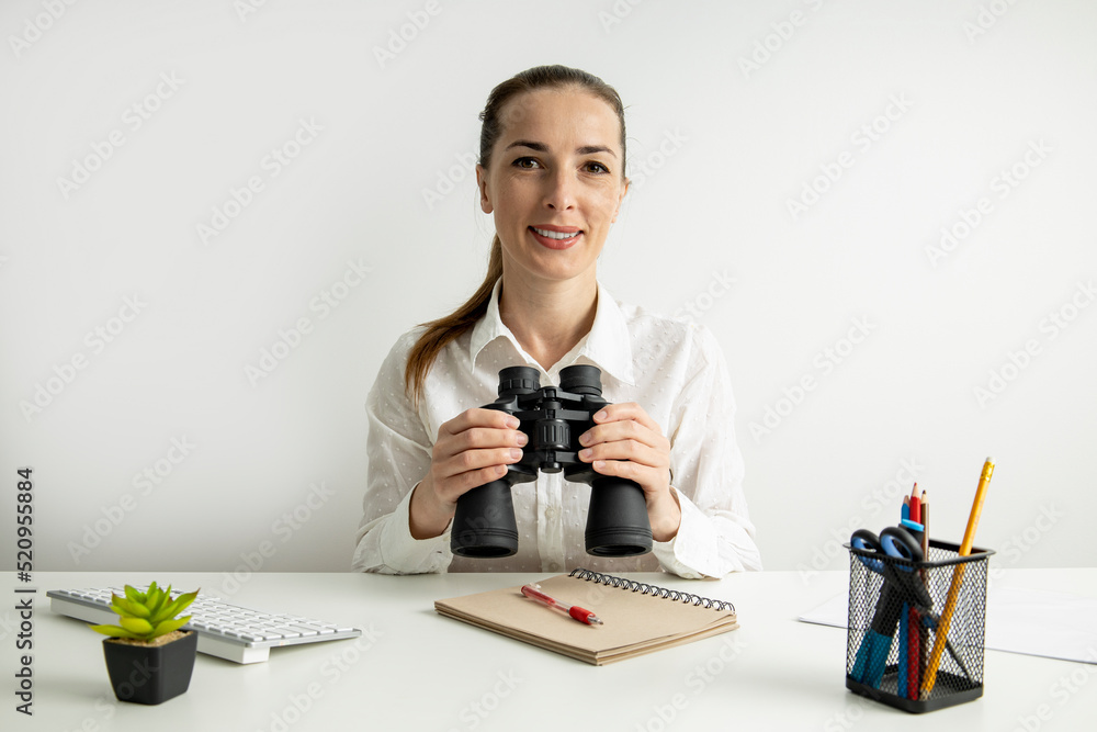 Smiling young woman with binoculars sitting at office desk on white background Smiling young woman with binoculars sitting at office desk on white background