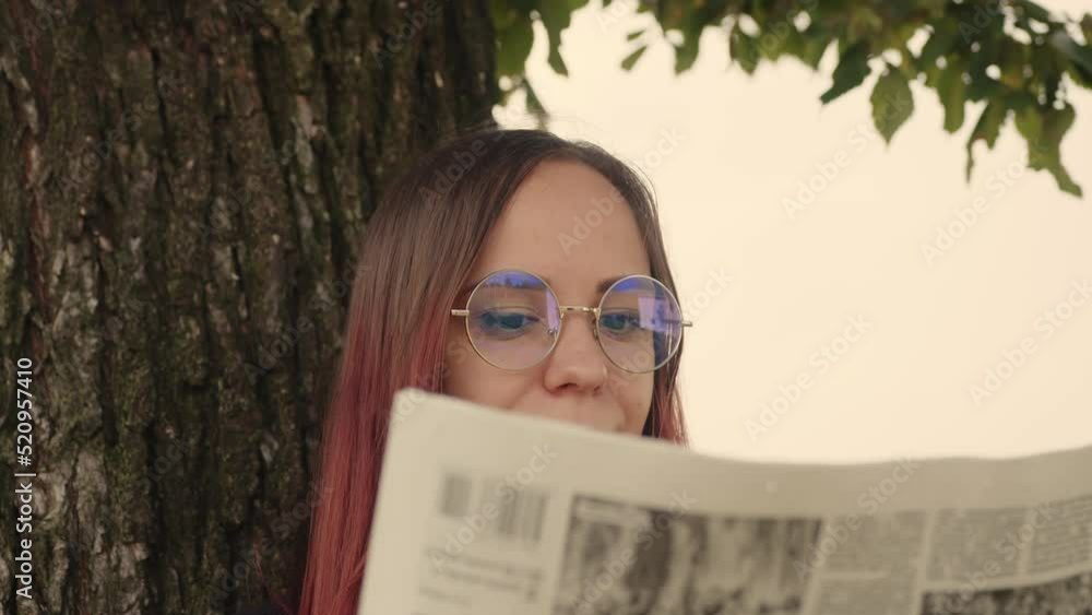Concentrated elegant lady reading newspaper in park. Focused young female entrepreneur with long dye hair in formal wear and eyeglasses standing near tree and reading newspaper with interest Concentrated elegant lady reading newspaper in park. Focused young female entrepreneur with long dye hair in formal wear and eyeglasses standing near tree and reading newspaper with interest